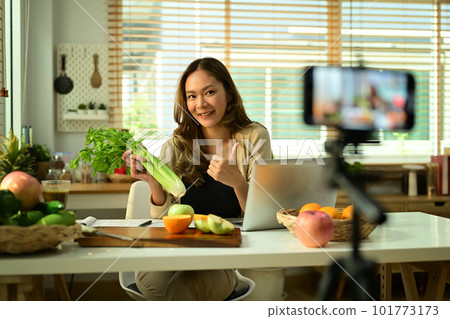 Confident female nutritionist holding celery, providing with online consultations or recording video content on smartphone Confident female nutritionist holding celery, providing with online consultations or recording video content on smartphone 101773173