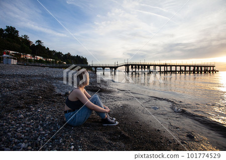 Young blonde woman sitting on pebble beach near sea on the background of pier and cloudy sky 101774529