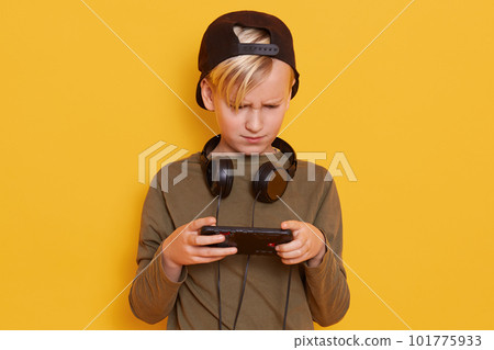 Studio shot of young caucasian little boy using mobile phone against yellow studio background, guy looks concentrated, holding his mobile phone, dresses green sweater and backwards black hat. Studio shot of young caucasian little boy using mobile phone against yellow studio background, guy looks concentrated, holding his mobile phone, dresses green sweater and backwards black hat. 101775933