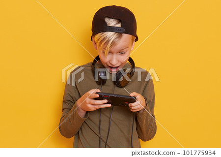 Close up portrait of little blond boy posing with mobile phone in hand isolated over yellow,, studio background, having astonished facial expression, keeps mouth opened, playing games concept. 101775934