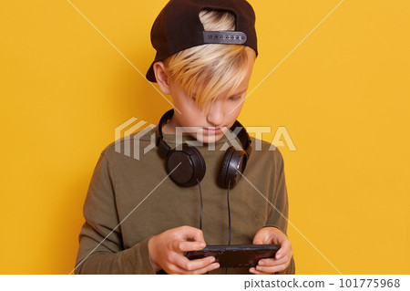 Close up of little boy using mobile phone while playing online games, cute male kid wearing gren hoody and black backwards hat keep headphones around neck. Modern technoloy concept, isolated on yellow Close up of little boy using mobile phone while playing online games, cute male kid wearing gren hoody and black backwards hat keep headphones around neck. Modern technoloy concept, isolated on yellow 101775968