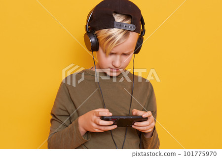Little child blond boy child playing mobile games on smartphone and using wireless Internet while listening to music via headphones, wearing stylish green hoody, looking down on his device's screen. 101775970