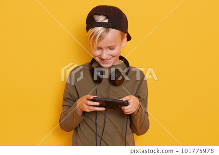 Close up portrait of handsome cute blonde little boy with gaming black headphones around his neck, looking directly at camera, posing isolated over yellow background, looks at his smartphone. Close up portrait of handsome cute blonde little boy with gaming black headphones around his neck, looking directly at camera, posing isolated over yellow background, looks at his smartphone. 101775978
