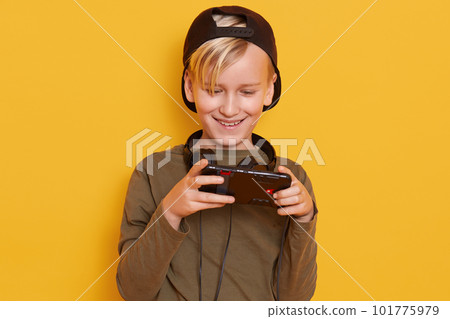 Horizontal shot of little boy wearing black cap and green hoody, posing with cell phone in hands, handsome child isolated over yellow studio background, fashionable kid playing online games. 101775979