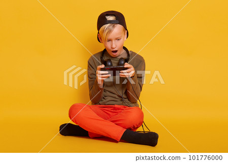Little boy sitting with smartphone in studio, guy wearing casually with headphones around neck, posing withopened mouth and looks excited, child with crossed legs, holding mobile phone in hands. 101776000