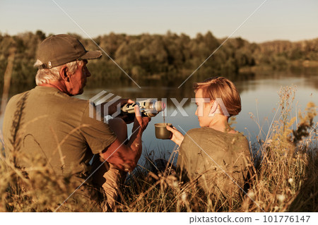 Two men, senior and little, sitting at bank of river and drinking hot coffee or tea from thermos flask talking, wearing casually. Beautiful view of lake and trees. Two men, senior and little, sitting at bank of river and drinking hot coffee or tea from thermos flask talking, wearing casually. Beautiful view of lake and trees. 101776147