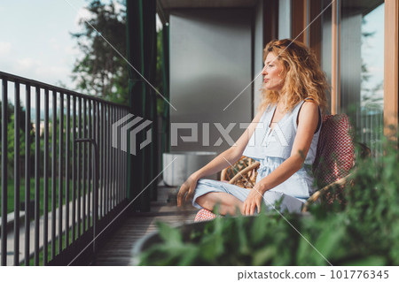 Side view of a woman relaxing on the balcony sitting in an armchair looking out in the nature  101776345