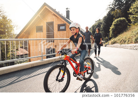 Smiling young girl riding a red bike while her parents walk behind her Smiling young girl riding a red bike while her parents walk behind her 101776356