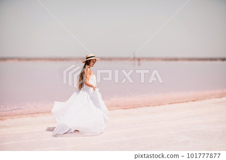 Woman in pink salt lake. She walks in a white long dress and hat along the salty white shore of the lake. Wanderlust photo for memory 101777877