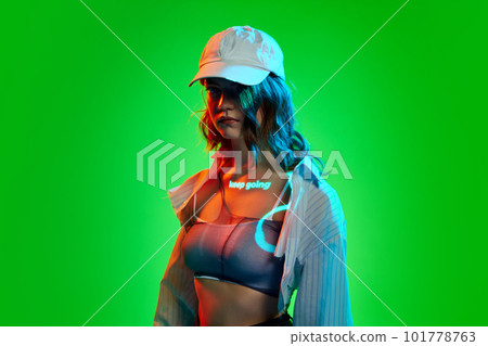 Creative portrait of young girl wearing cap with digital neon filter lights with inscryption on body on bright green mode background. Keep going 101778763