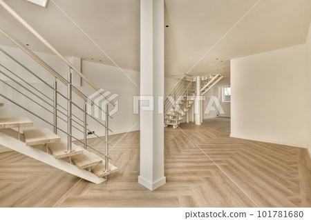 an empty room with wood flooring and staircases in the middle part of the room there is a white wall on the right 101781680