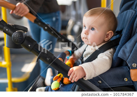 A cute little baby of 10 months is sitting in a stroller. A child is fastened with seat belts in stroller rides a passenger bus with her mother. Concept of child safety. 101784367