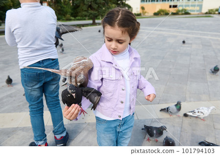 Caucasian little girl looking at a pigeon sitting on her arm, while feeding flock of flying doves in city park. The concept of a happy carefree childhood and instilling love for nature and animals 101794103