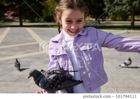 Happy overjoyed Caucasian little child girl rejoices at a pigeon sitting on her arm, smiling, expressing happiness while feeding flock of flying birds in the city park on a sunny spring day Happy overjoyed Caucasian little child girl rejoices at a pigeon sitting on her arm, smiling, expressing happiness while feeding flock of flying birds in the city park on a sunny spring day 101794111