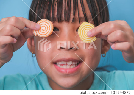 Smiling cute little girl covering her eyes with two gelatinous sweets isolated on blue background.  Children eat sugary sweets, causing loss teeth or tooth decay and unhealthy oral care. 101794872
