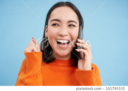 Cheerful asian girl talks on mobile phone, speaks with someone on telephone, stands over blue background Cheerful asian girl talks on mobile phone, speaks with someone on telephone, stands over blue background 101795335