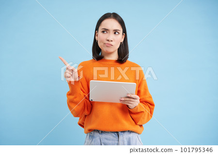 Portrait of korean woman with digital tablet, pointing at upper left corner with confused, frowning face, standing over blue background 101795346