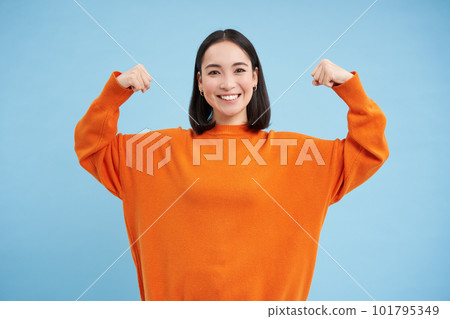 Portrait of cute korean woman flexing her biceps, shows strong arms, muscles and smiles, concept of healthy and fit people, blue background Portrait of cute korean woman flexing her biceps, shows strong arms, muscles and smiles, concept of healthy and fit people, blue background 101795349