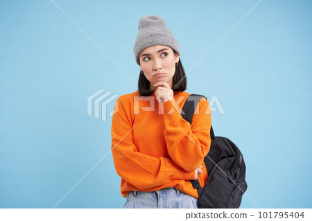 Thinking woman in hat, holds backpack, ponders, thinking or making decision, standing over blue studio background 101795404