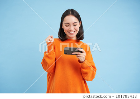 Happy korean girl winning on mobile phone video game, holding cellphone and celebrating, achieve goal in app, standing over blue background 101795418