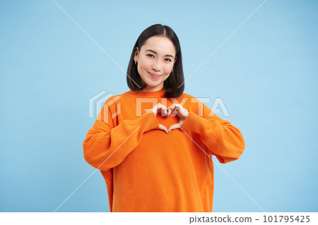 Lovely asian woman, korean girl shows heart gesture and smiles with care and tenderness, stands in orange sweatshirt against blue background Lovely asian woman, korean girl shows heart gesture and smiles with care and tenderness, stands in orange sweatshirt against blue background 101795425