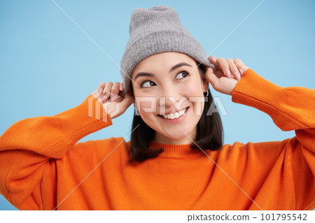 Close up portrait of smiling asian woman in warm hat, looking happy and cute at camera, has clear natural skin, stands over blue background 101795542