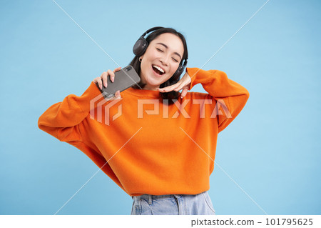 Beautiful happy korean woman in headphones, listening music on mobile phone in earphones, wearing orange sweatshirt, blue background 101795625