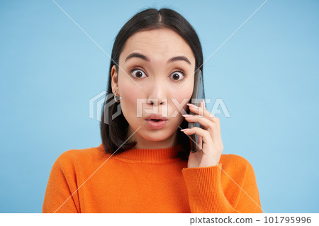 Technology concept. Asian girl answers phone call and looks surprised at camera, stands over blue background Technology concept. Asian girl answers phone call and looks surprised at camera, stands over blue background 101795996