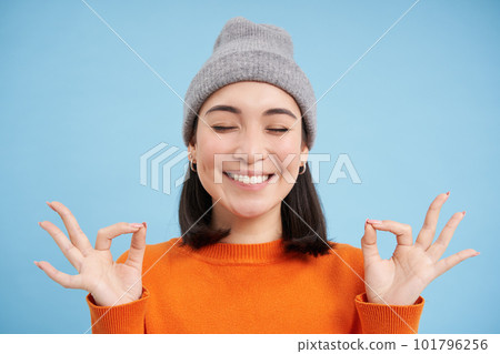 Sounds mind in healthy body. Smiling calm and relaxed asian girl in beanie, shows zen, relaxation gesture, meditating, standing over blue background 101796256