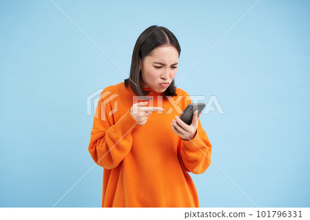 Annoyed girl with phone, points at mobile screen with frowning angry face, standing disappointed over blue background 101796331