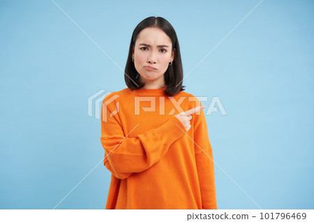 Upset brunette girl 25 years old, points right at banner, copy space on blue background, frownd with disappointed face expression Upset brunette girl 25 years old, points right at banner, copy space on blue background, frownd with disappointed face expression 101796469