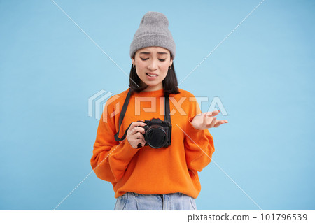 Portrait of asian woman in hat, holding digital camera with confused face, unprofessional photographer doesnt know how to take pictures on digicam, blue background Portrait of asian woman in hat, holding digital camera with confused face, unprofessional photographer doesnt know how to take pictures on digicam, blue background 101796539