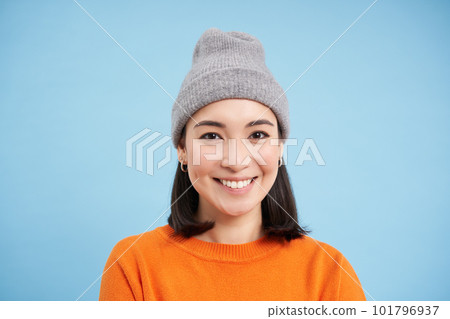 Close up portrait of smiling asian woman in warm hat, looking happy and cute at camera, has clear natural skin, stands over blue background 101796937