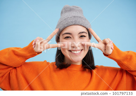 Close up portrait of cute korean girl in beanie, shows peace, vsign gesture, positive vibe, smiling and laughing, posing over blue background Close up portrait of cute korean girl in beanie, shows peace, vsign gesture, positive vibe, smiling and laughing, posing over blue background 101797133