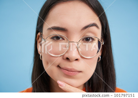 Close up portrait of asian woman looks intrigued, wears glasses, squints thoughtful, thinking, making assumption, standing over blue background Close up portrait of asian woman looks intrigued, wears glasses, squints thoughtful, thinking, making assumption, standing over blue background 101797151