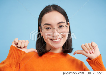 Portrait of cute korean woman in glasses, pointing fingers at camera self-assured, smiles with confidence, blue background 101797223