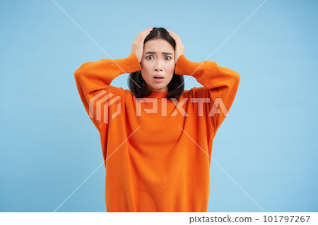 Shocked and anxious asian woman, holds hands on head, looks worried, doesnt know what to do, looks complicated, stands over blue background Shocked and anxious asian woman, holds hands on head, looks worried, doesnt know what to do, looks complicated, stands over blue background 101797267