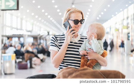 Mother talking on mobile phone while traveling with child, holding his infant baby boy at airport terminal waiting to board a plane. Travel with kids concept 101798591