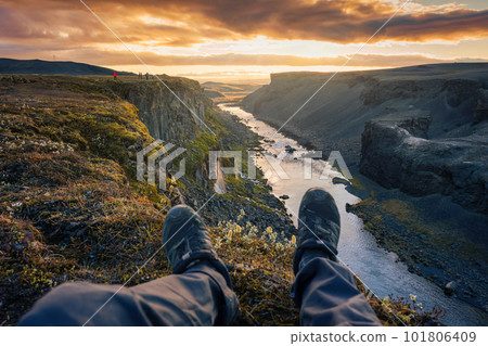 Legs of hiker man sitting on edge of mountain with waterfall flowing in canyon on Icelandic Highlands in the evening at Sigoldugljufur 101806409