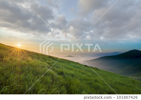 Mountain landscape in nice weather at sunrise. Green grassy steep hill, foggy valley and distant mountains under bright blue sky with lit by raising sun white clouds. Beauty of nature concept. 101807426