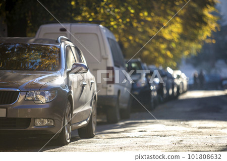 Long row of cars parked along roadside on sunny autumn day on blurred green golden foliage bokeh background. Transportation, modern city lifestyle, vehicles parking problems concept. 101808632