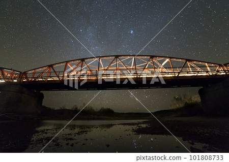Illuminated metal bridge on concrete supports reflected in water on dark starry sky with Milky Way constellation background. Night photography concept. 101808733