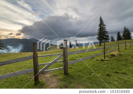 Green grassy valley slope with wooden fence and gate on woody foggy mountain under blue cloudy sky. 101816880