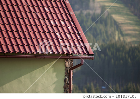 Detail of new modern house cottage corner with stucco walls decorated with natural stones, red shingled roof and rain gutter pipe system on blurred spruce forest copy space background. 101817103