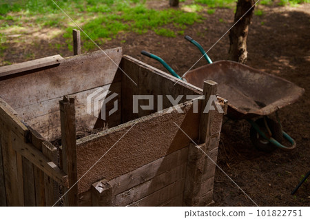 Close-up of wooden compost bin near empty wheelbarrow in the country house backyard. Composting and eco farming concept. Eco farming and agricultural hobby concept. Zero waste concept. Close-up of wooden compost bin near empty wheelbarrow in the country house backyard. Composting and eco farming concept. Eco farming and agricultural hobby concept. Zero waste concept. 101822751