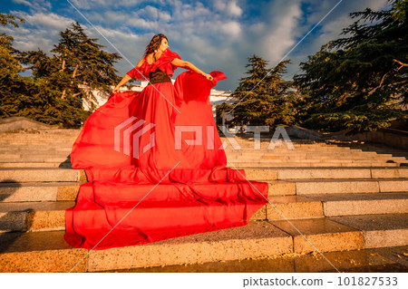 Sunrise red dress. A woman in a long red dress against the backdrop of sunrise, bright golden light of the sun's rays. The concept of femininity, harmony. 101827533