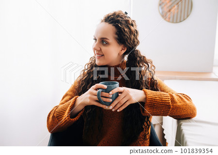 Young smiling pretty woman holding cup drinking warm tea or coffee relaxing dreaming at home. Happy 101839354