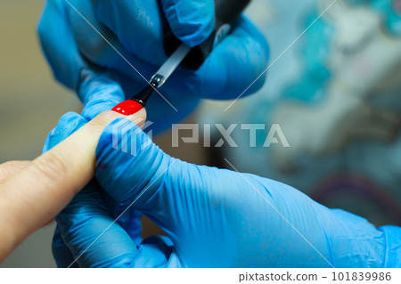 A manicurist in blue gloves covers the nails with a long-lasting red gel polish. 101839986