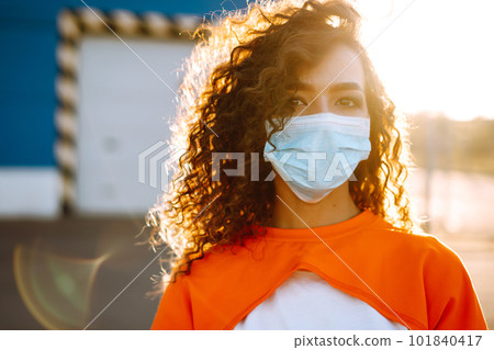 Young woman with medical face mask at sunset. Curly woman posing during quarantine of coronavirus pandemic. Young woman with medical face mask at sunset. Curly woman posing during quarantine of coronavirus pandemic. 101840417