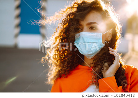 Young woman with medical face mask at sunset. Curly woman posing during quarantine of coronavirus pandemic. 101840418